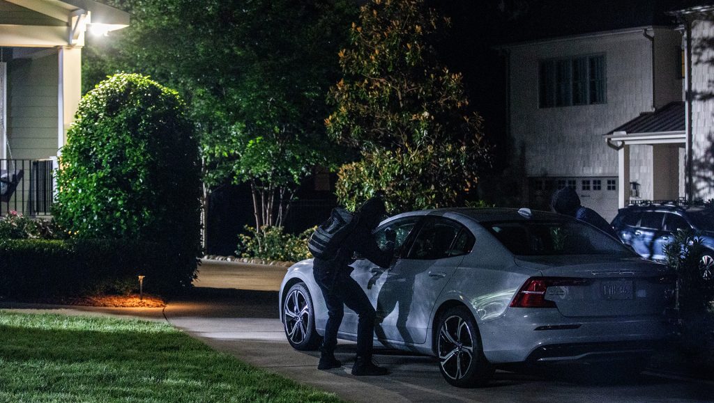 Man dressed in black standing outside a white sedan in a home’s driveway at night