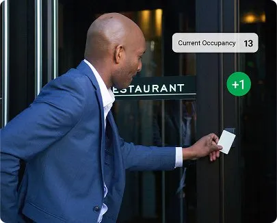 Person in a blue suit tapping a white card on a black access control box at a restaurant door, with overlaid text showing 'Current occupancy 13' and a green '+1' circle