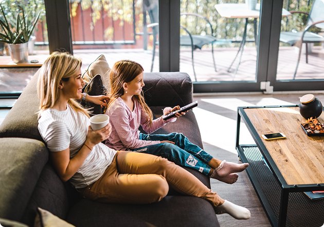 Woman sitting on a couch next to a young girl holding a remote, with modern living room furniture in the background